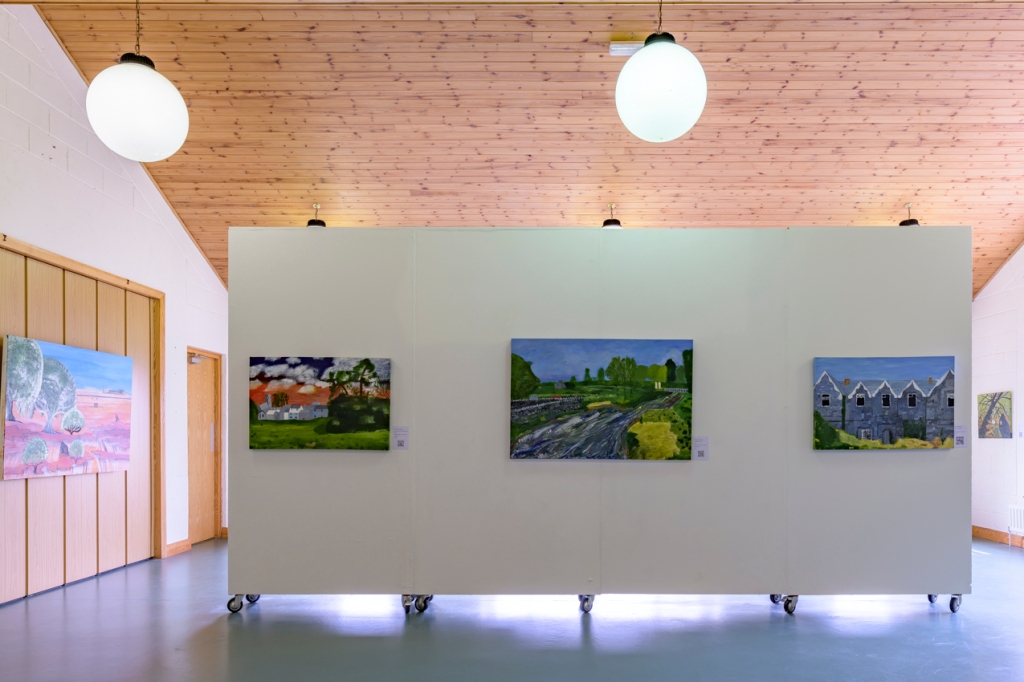Installation shot of exhibition. In the centre three paintings hang on a white wall in a high ceilinged room. There are other paintings hanging on the wall in the background.