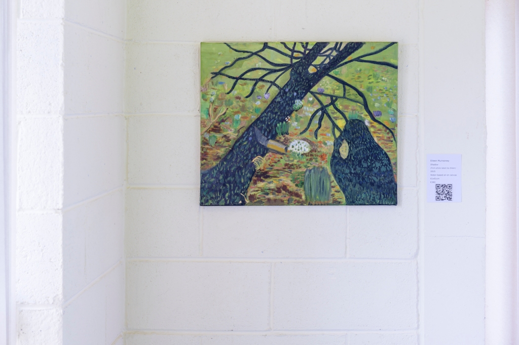 This is a photo of a painting by Eileen Mulrooney of a shadow of a tree on grass and flowers. The painting is hung on a white breeze block wall in a corner. There is a QR code hung on the right hand side of the painting.